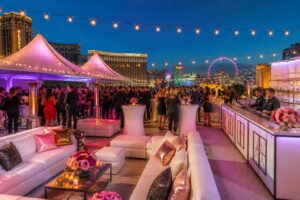 An outdoor evening party in Las Vegas with tents, lounge furniture, string lights, a bar, and guests enjoying the event with the city skyline in the background.